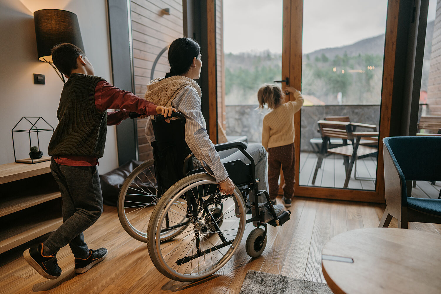 Zwei Kinder und eine Frau im Rollstuhl zusammen in einer Wohnung, ein Kind öffnet die Balkontür, das zweite schiebt den Rollstuhl