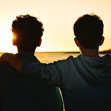 Zwei Jungs gucken in einen Sonneuntergang am Meer, der eine hat den Arm um den anderen gelegt.