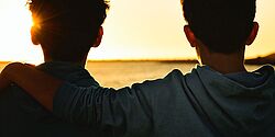 Zwei Jungs gucken in einen Sonneuntergang am Meer, der eine hat den Arm um den anderen gelegt.