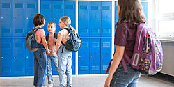 Vier Kinder stehen im Flur einer Schule. Ein Kind steht abseits der Gruppe und guckt traurig in ihre Richtung.