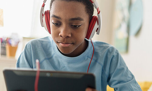 Jugendlicher sitzt entspannt auf einem Bett und schaut konzentriert auf ein Tablet, während er rote Kopfhörer trägt. 