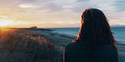 Eine Frau ist bis zum Rücken zu sehen, welche dem Foto abgewandt in Richtung der Sonne blickt, die gerade zwischen Strand und Meer im Hintergrund untergeht.